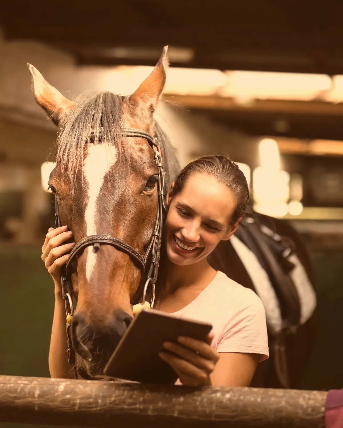 A rider smiling at her tablet beside her horse in the stable